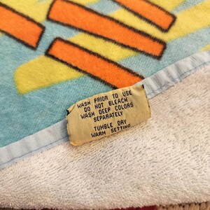 May include: A colorful beach towel with a geometric design in orange, yellow, and black on a light blue background. The towel has a white border and a care instruction tag. The tag reads "WASH PRIOR TO USE" and "TUMBLE DRY".