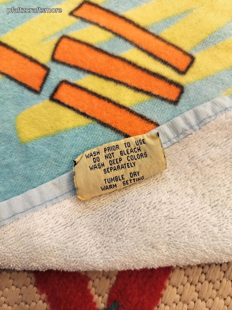 May include: A colorful beach towel with a geometric design in orange, yellow, and black on a light blue background. The towel has a white border and a care instruction tag. The tag reads "WASH PRIOR TO USE" and "TUMBLE DRY".