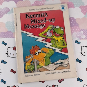 Puede incluir: Un libro infantil vintage titulado "Kermit's Mixed-up Message" con Kermit la Rana y otros personajes de los Muppets. La portada es roja, azul y amarilla, con ilustraciones de los personajes usando teléfonos. El libro es de Joanne Barkan e ilustrado por Lauren Attinello.