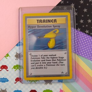 Può includere: Una carta da gioco di scambio Pokémon gialla e blu che presenta una carta allenatore chiamata "Hyper Devolution Spray". La carta mostra un flacone spray giallo e blu con un ugello rosso. Il testo della carta recita: "Scegli 1 dei tuoi Pokémon evoluti. Prendi la carta di Evoluzione di Fase più alta di quel Pokémon e mettila nella tua mano. (Non puoi far evolvere un Pokémon nel turno in cui lo fai evolvere all'indietro.)"