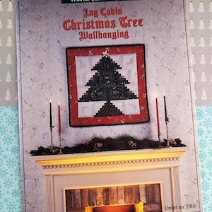 May include: A book cover for a "Log Cabin Christmas Tree Wallhanging" quilt pattern. The cover features a finished quilt with a black and red Christmas tree design, a star, and the text "From the Quilt in a Day Series."