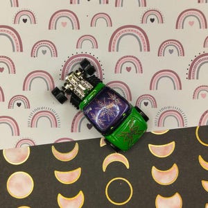 Könnte beinhalten: Ein grünes und violettes Spielzeugauto mit Flammen, die auf die Seite gemalt sind, auf einem Hintergrund aus rosa und grauen Regenbögen und einem schwarzen Hintergrund mit goldenen und rosafarbenen Monden.