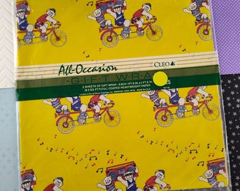 Puede incluir: Dos hojas de papel de regalo amarillo con un patrón repetido de osos de dibujos animados montando una bicicleta tándem con una radio. El texto "All-Occasion Gift Wrap" está impreso en la parte inferior de la hoja. CLEO 25 está impreso en un círculo en el lado derecho de la hoja.