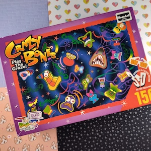 May include: A 150-piece jigsaw puzzle box featuring a colorful illustration of cartoon characters with a purple, orange, and black background. The box has the words "Crazy Bones" and "Play The Craze!" on the front.