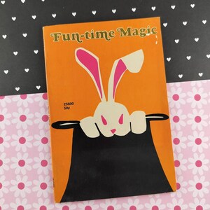 Puede incluir: Una portada de libro naranja con el título "Fun-time Magic" presenta un conejo blanco con orejas rosas saliendo de un sombrero de copa negro. El fondo es negro con corazones blancos.