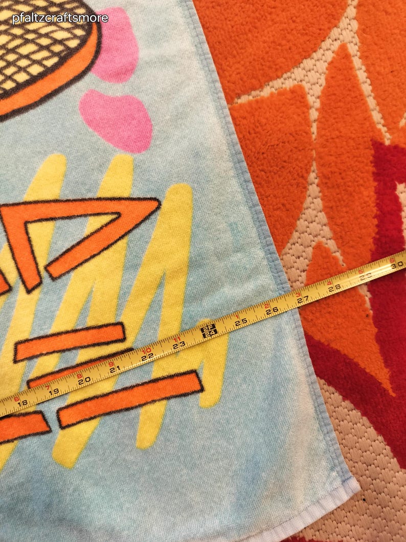 May include: A light blue beach towel with a tennis racket design and abstract shapes in orange, yellow, and pink. The towel measures approximately 29 inches in length.
