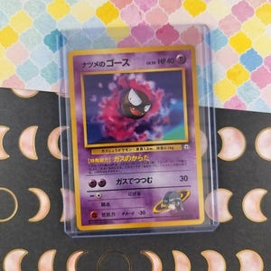 Può includere: Una carta collezionabile di Pokémon con un personaggio Gastly viola e nero con testo giapponese. La carta è in una custodia protettiva. Lo sfondo è un motivo colorato di forme gialle, rosa e bianche. Peso 0,1 kg.