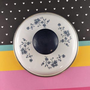 May include: A round, white lid with a dark blue knob. The lid features a floral pattern in shades of blue and is trimmed with silver. The background is a mix of colors and patterns.
