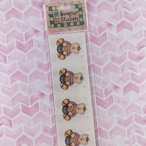 May include: A vertical strip of four identical stickers in clear packaging. Each sticker features a light brown teddy bear wearing a red and white striped shirt and a small blue hat, sitting beside a yellow honey pot. The header reads Designer Stickers.