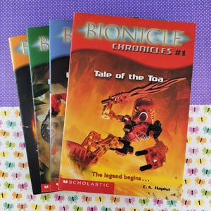 Op de afbeelding: Een stapel Bionicle Chronicles #1 boeken. De omslag toont een rode robot met de titel "Tale of the Toa" en de tekst "The legend begins..." door C.A. Hapka. De boeken liggen op een kleurrijk oppervlak met vlinderpatroon.