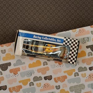 Puede incluir: Una caja blanca con un patrón de cuadros negros y blancos en el lateral. La caja contiene un pequeño coche de juguete amarillo y negro con las palabras "Racing Collectables, Inc." impresas en la caja.