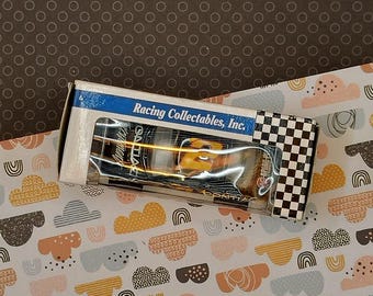 Op de afbeelding: Een witte doos met een zwart-wit geruit patroon aan de zijkant. De doos bevat een kleine, gele en zwarte speelgoedauto met de tekst "Racing Collectables, Inc." erop gedrukt.