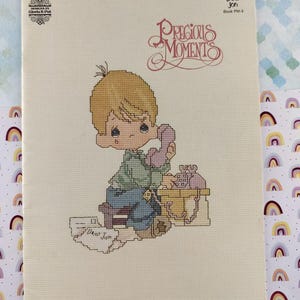 May include: A cross-stitch pattern featuring a Precious Moments design. The image shows a young boy with blonde hair, holding a pink telephone. The text "Dear Jon" is visible. The design is on a cream-colored background.