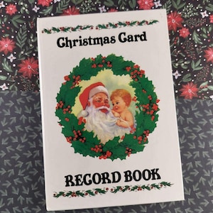 May include: A white Christmas Card Record Book with a festive design. The cover features Santa Claus and a cherub inside a holly wreath. The words "Christmas Card" and "Record Book" are printed in black. The background is a floral pattern.