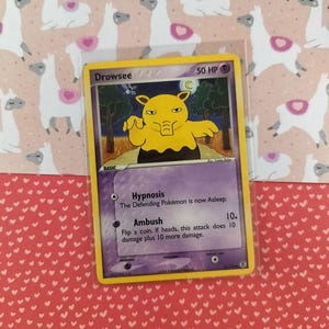 Puede incluir: Una carta coleccionable de Pokémon Drowsee en una funda protectora. La carta presenta una criatura amarilla con un borde morado y el texto "Drowsee" y "50 HP". El fondo es un patrón de llamas rosas y blancas.