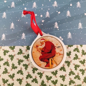 Puede incluir: Un adorno navideño blanco con un diseño circular que presenta a Papá Noel con un traje rojo, sentado en un taburete. El adorno tiene una cinta roja para colgar. El fondo es un patrón festivo con acebo y árboles de Navidad.