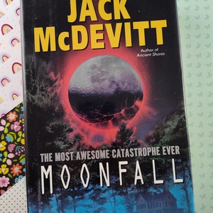 Può includere: Copertina del libro "Moonfall" di Jack McDevitt, autore di "Ancient Shores". La copertina presenta uno sfondo blu scuro con una grande luna rossastra al centro, circondata da un anello di luce. Il titolo "Moonfall" è scritto in lettere bianche, e il nome dell'autore è scritto in lettere gialle.