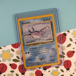 May include: A Mantine Pokémon trading card in a protective sleeve. The card features a blue and purple manta ray illustration with the name "Mantine" at the top. The card has a yellow border and is on a patterned background.