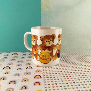 May include: White ceramic mug with a design of three cartoon teddy bears, each with a red bow. The bears are brown with black outlines. A yellow honey pot with the word "HONEY" is also featured. The mug has a white handle.