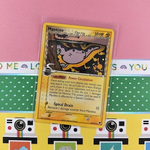 May include: A Mantine Pokémon trading card in a protective sleeve. The card features a cartoon illustration of Mantine with a yellow border and text describing its abilities. The background is pink with decorative elements.