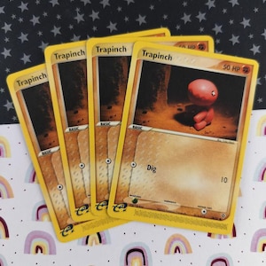 Może przedstawiać: Cztery karty kolekcjonerskie Pokémon Trapinch. Karty mają żółtą obwódkę i przedstawiają czerwoną istotę z tekstem "Dig" i liczbą "10". Tło jest ciemne i ziemiste. Karty leżą na wzorzystej powierzchni.