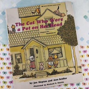 May include: A white ceramic mug with a black handle and the text "But First, Coffee" in a bold, black font. The mug is set against a blurred background, suggesting a cozy setting.