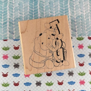 May include: A wooden stamp featuring an elephant with the letters "T U" and a telephone receiver. The stamp has a light brown wooden base and a black ink design. The background is a patterned fabric with blue, green, and pink shapes.