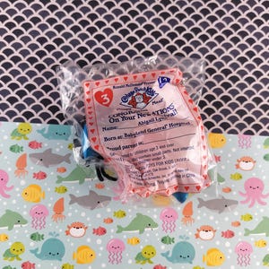 May include: A sealed Happy Meal toy from McDonald's, featuring a Cabbage Patch Kids theme. The packaging is pink and red, with text that reads "CONGRATULATIONS On Your New Arrival!" and the name "Abigail Lynival!"