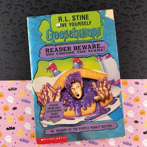 以下が含まれることがあります： R.L. Stineの「Beware of the Purple Peanut Butter」というタイトルのヴィンテージグースバンプス本。表紙には、紫色のピーナッツバターの中にいる人の漫画イラストが描かれています。本はピンク色のハロウィーンをテーマにした背景に置かれています。
