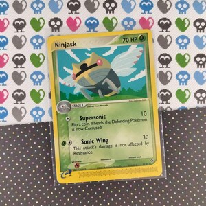 Pode incluir: Um cartão colecionável Pokémon Ninjask com borda amarela, apresentando uma criatura parecida com um inseto com asas. O cartão exibe o texto "Ninjask" e "70 HP", juntamente com descrições de ataque. O fundo é um padrão de caveiras, corações e formas azuis e verdes.