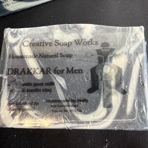 Puede incluir: Una pastilla de jabón natural casero, envuelta en plástico transparente. El jabón tiene un diseño veteado en blanco y negro. El texto del embalaje dice "Creative Soap Works", "DRAKKAR for Men" y "with goat milk & kaolin clay."