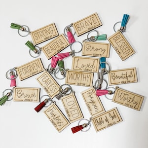 May include: A collection of wooden keychains with inspirational words and bible verses. The keychains are decorated with colorful tassels and feature phrases like "Brave", "Strong", "Loved", "Worthy", "Forgiven", "Fearless", "Beautiful", "Blessed", and "Child of God".