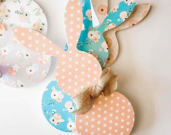 Può includere: Decorazioni pasquali a forma di coniglietto in legno. I coniglietti presentano una varietà di motivi, tra cui pois e disegni floreali. Un coniglietto ha un fiocco di juta. La tavolozza dei colori comprende pesca, blu e bianco.
