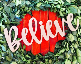 Può includere: Una ghirlanda di bosso verde con la parola "Believe" in lettere bianche su uno sfondo rosso.