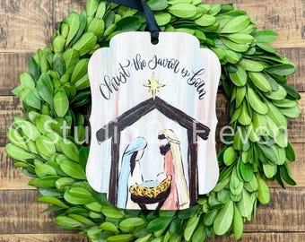 May include: A wooden Christmas ornament with a watercolor painting of the nativity scene. The ornament is white with a light blue, pink, and yellow watercolor background. The text "Christ the Savior is Born" is written in black calligraphy on the ornament.