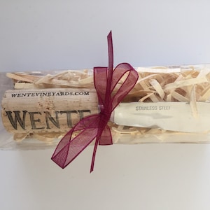 May include: A clear plastic box with wood shavings and a burgundy ribbon contains a silver butter knife and a wine cork with the text "WENTE VINEYARDS.COM" and "WENTE" printed on it.