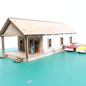 May include: A wooden model of a small house with a porch and two doors. The house is painted white and has a brown roof. There are two small toy cars parked in front of the house.
