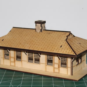 May include: A wooden model of a train station with a brown roof and a chimney. The station has four windows and a door. The model is sitting on a green cutting mat.
