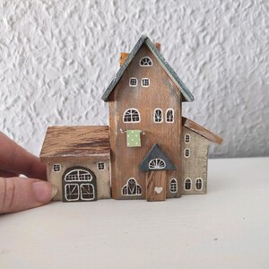 May include: A miniature wooden house with a brown roof, white trim, and a green door. The house has a small window on the side and a larger window on the front. The house is painted in a rustic style.
