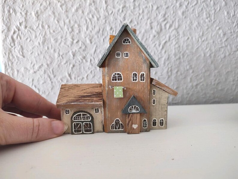 May include: A miniature wooden house with a brown roof, white trim, and a green door. The house has a small window on the side and a larger window on the front. The house is painted in a rustic style.