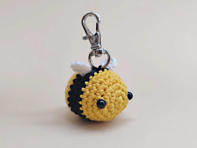 May include: A crocheted yellow and black bee keychain with white wings and black bead eyes. The bee is attached to a silver metal key ring.
