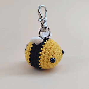 May include: A crocheted bee keychain with black and yellow stripes, white wings, and black bead eyes. The keychain has a silver metal loop.