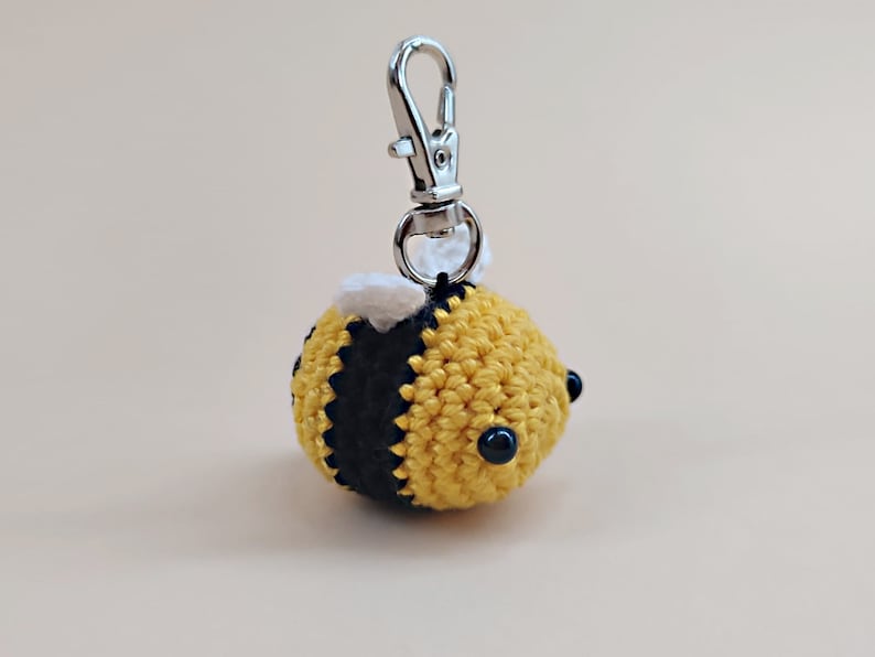 May include: A crocheted bee keychain with black and yellow stripes, white wings, and black bead eyes. The keychain has a silver metal loop.