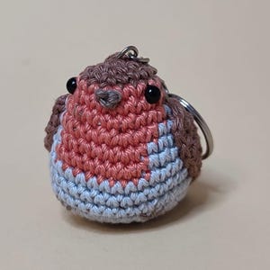 May include: A handmade crochet keychain in the shape of a robin bird. The bird features a red breast, blue belly, and brown head and wings. The keychain has black eyes and a silver metal ring.