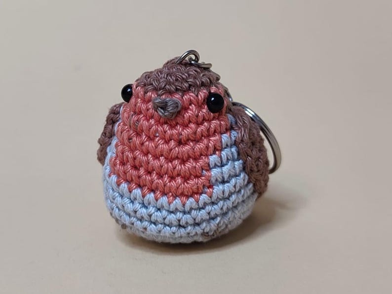 May include: A handmade crochet keychain in the shape of a robin bird. The bird features a red breast, blue belly, and brown head and wings. The keychain has black eyes and a silver metal ring.