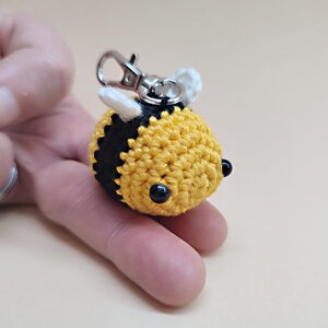 May include: A crocheted bee keychain with black and yellow stripes, white wings, and black eyes. The bee is attached to a silver key ring.