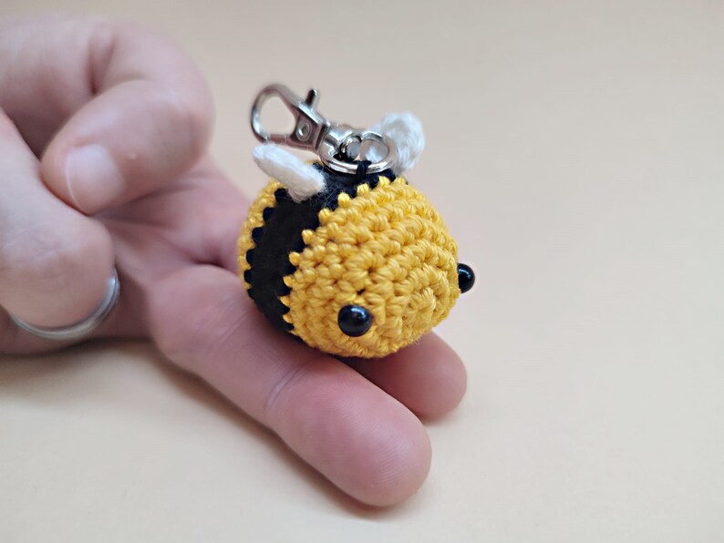 May include: A crocheted bee keychain with black and yellow stripes, white wings, and black eyes. The bee is attached to a silver key ring.