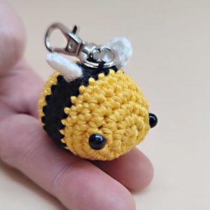 May include: A crocheted bee keychain with black and yellow stripes, white wings, and black bead eyes. The bee is attached to a silver metal key ring.