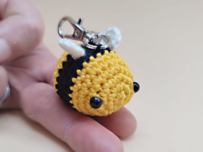 May include: A crocheted bee keychain with black and yellow stripes, white wings, and black bead eyes. The bee is attached to a silver metal key ring.