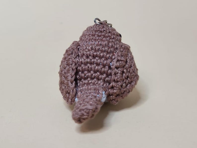 May include: A handmade, crocheted bird keychain in shades of brown. The bird has a rounded body, wings, and a small tail. A silver-colored metal ring is attached to the top for hanging. The bird is made with a textured stitch.